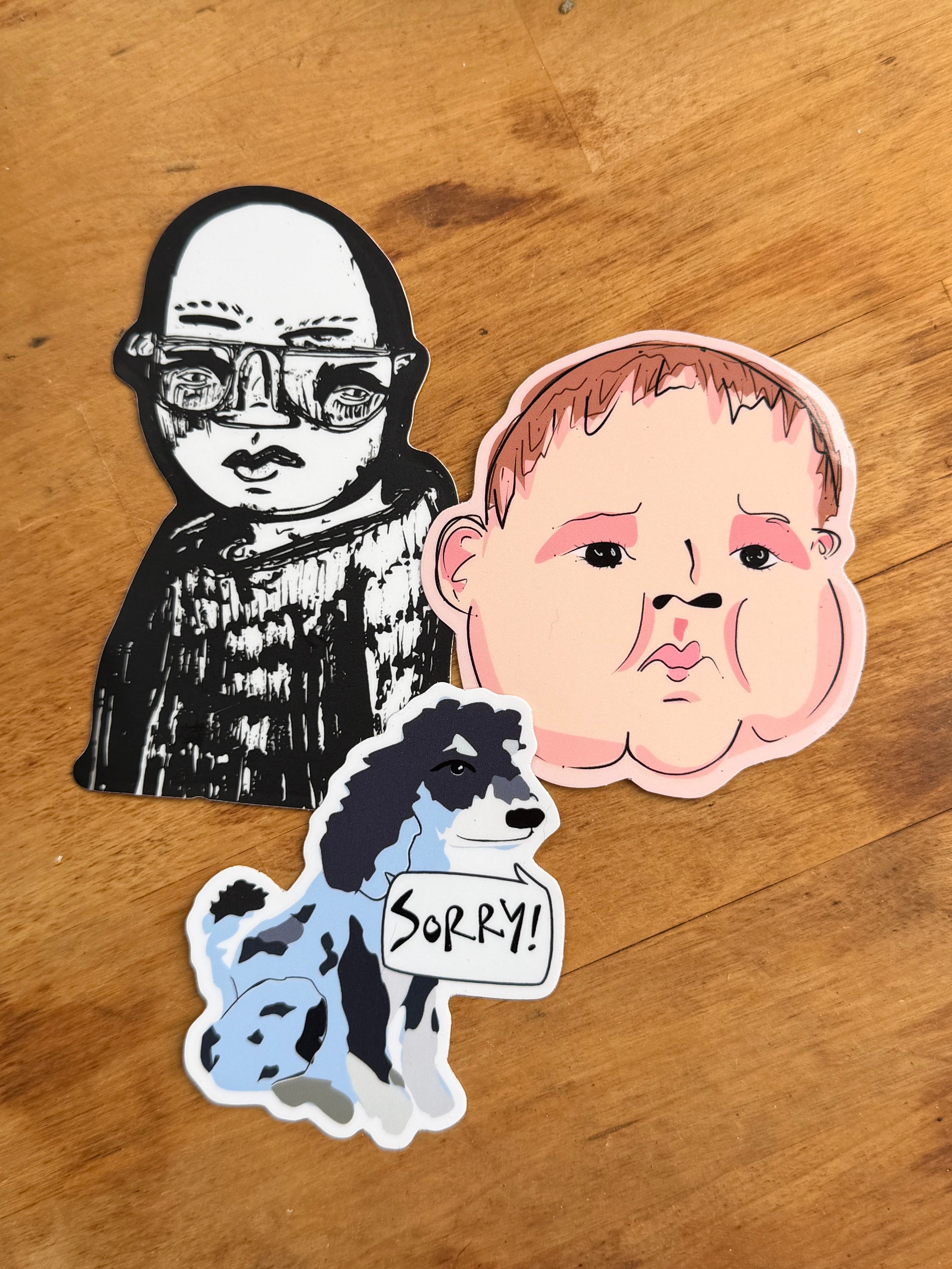 Three cartoon-style stickers on a wooden tabletop: a black-and-white illustrated figure with glasses, a pink-toned face sticker, and a blue-and-white dog holding a sign that says 'Sorry!'.